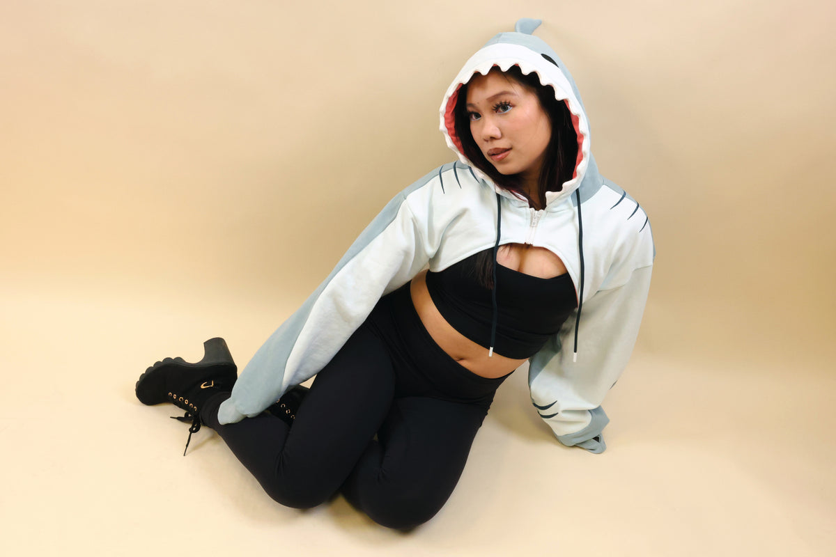 Model kneeling in the Nyahallo Shark Shrug, featuring a light blue shark hood with white teeth, pink lining, gill details, and oversized sleeves over a black outfit.