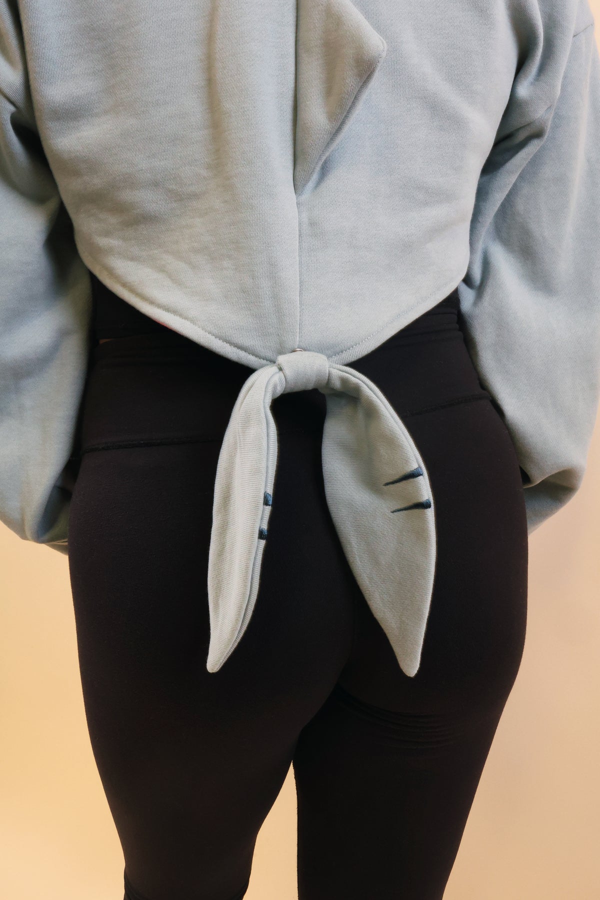 Close-up of the back of the Nyahallo Shark Shrug, highlighting its light blue fabric, cropped hem, and dangling shark tail with embroidered details.
