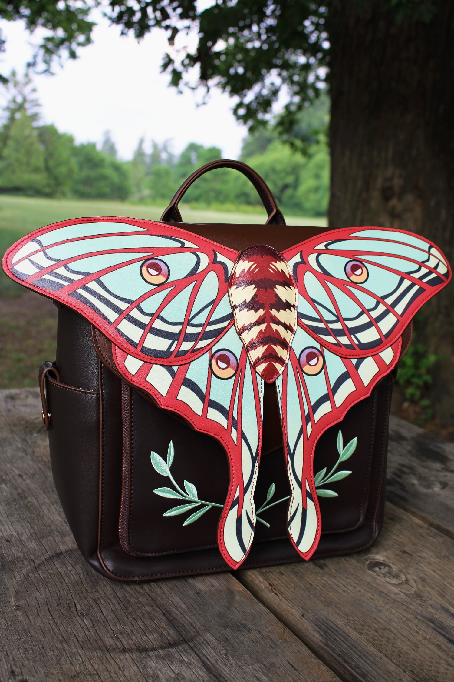 Front view of spanish moon moth backpack on a wooden table outdoors, showcasing large detailed moth wing flap and leaf embroidery.
