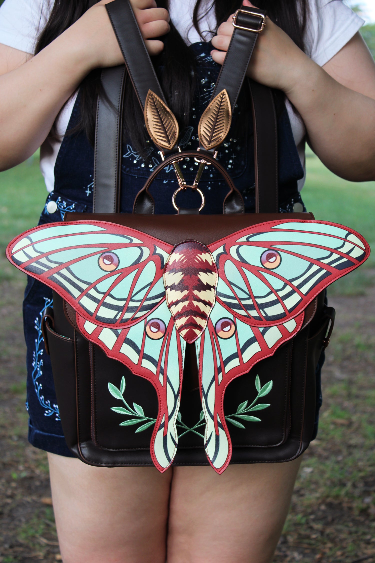 A striking backpack featuring an oversized spanish moon moth wing design in rich burgundy, teal, and cream tones. Styled with metallic gold leaf-shaped hardware and twin adjustable straps, this piece is shown being held outdoors to highlight its elegant silhouette and artisan craftsmanship. Perfect for statement styling and nature lovers alike.