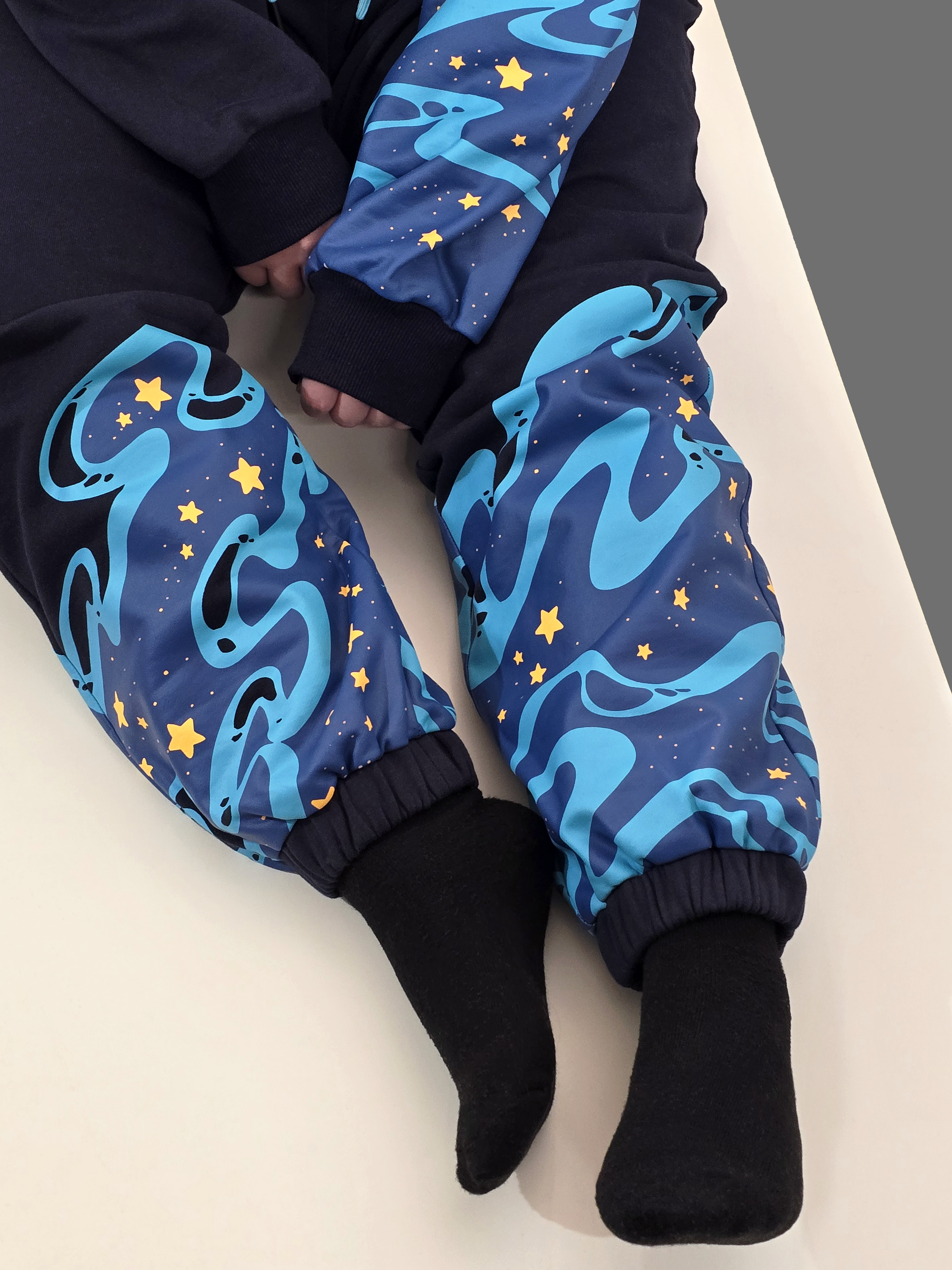 Navy Stellar Tide sweatpants with blue galaxy wave patterns and scattered yellow stars shown in a close-up cozy indoor photo.
