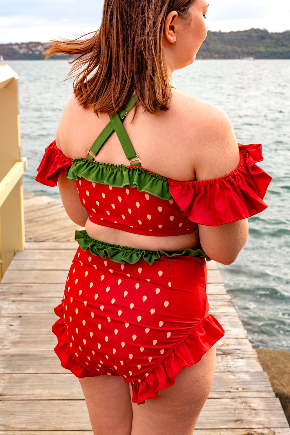 Back view of a whimsical strawberry-inspired swimsuit with red ruffles, strawberry print, and green strap details by the water.