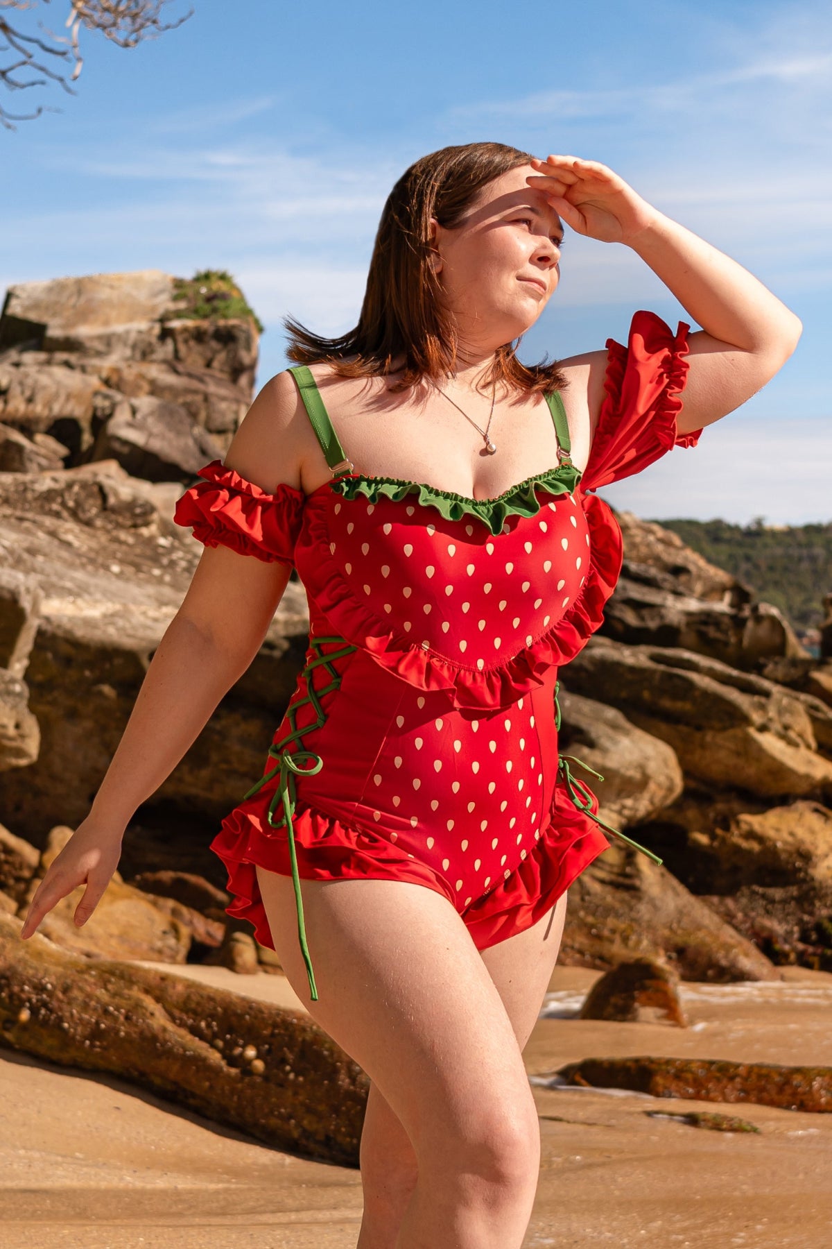 Model wearing the Nyahallo Strawberry One-Piece Swimsuit featuring a strawberry print, off-shoulder ruffle sleeves, leafy green trim, adjustable side lace-up ties, and a ruffle hem on a rocky beach.