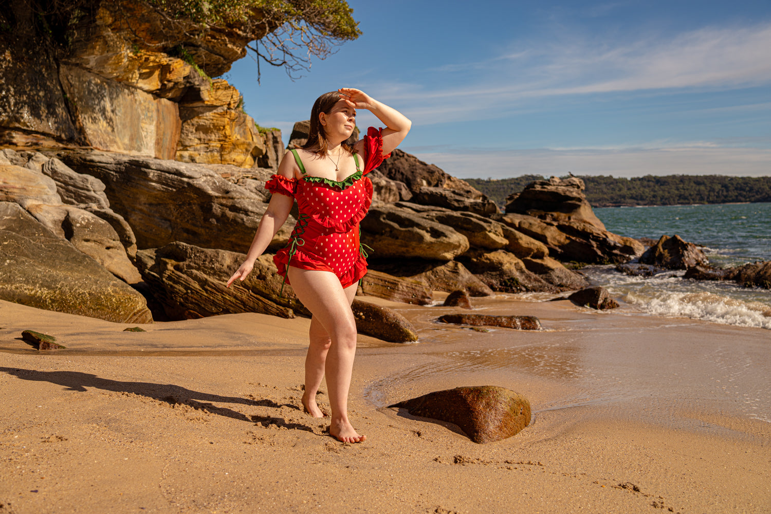 Model wearing Nyahallo strawberry swimsuit on a sunny beach with rocky shoreline