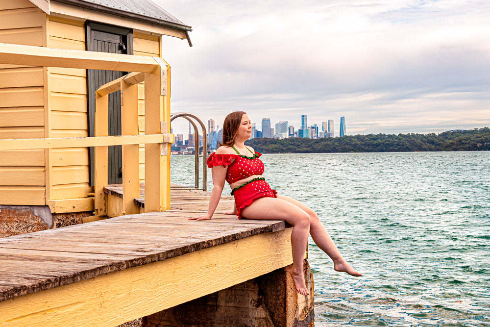 Model wearing Nyahallo strawberry swimsuit sitting on a dock with city skyline in background