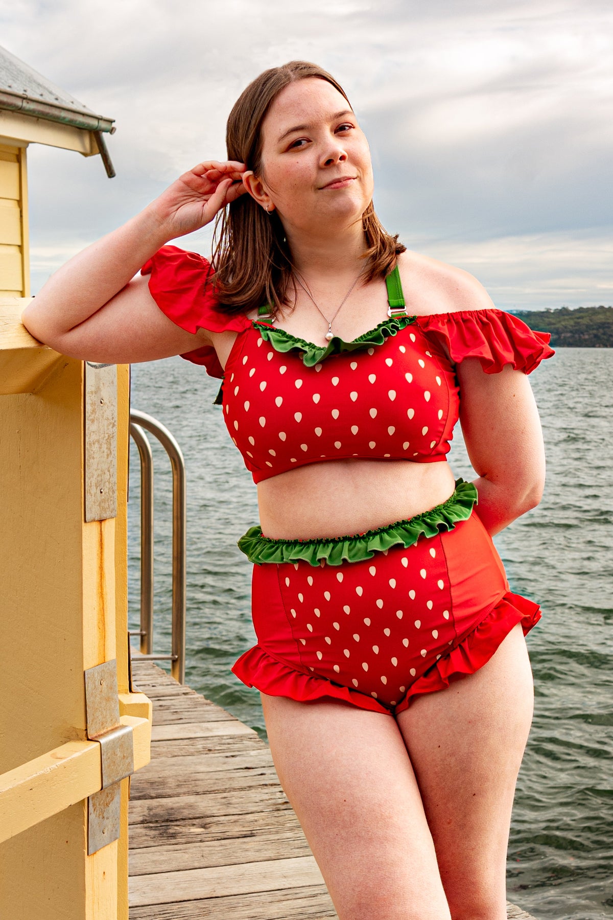 Model wearing the Nyahallo Strawberry Swimsuit with an off-shoulder strawberry-print top and high-waisted ruffle bottoms on a lakeside dock.