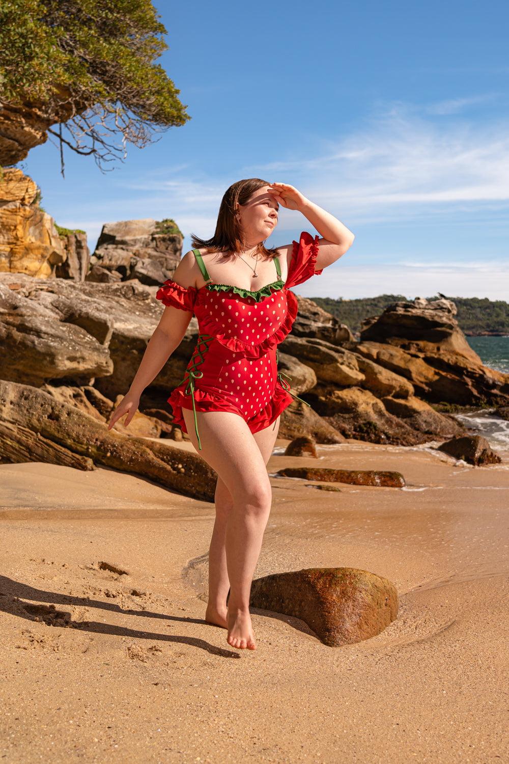 Model wearing a red strawberry-themed one-piece swimsuit with green ribbon details and off-shoulder ruffles, walking on a sunny beach with rocky cliffs in the background.