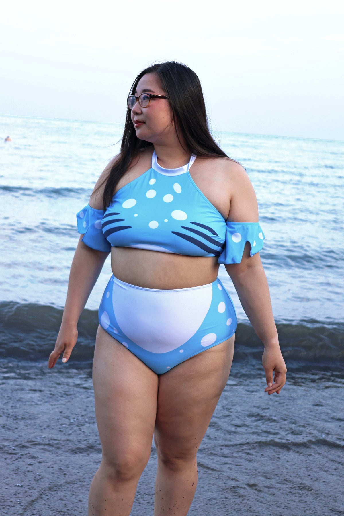 Model wearing the blue and white Whale Shark two-piece swimsuit with spotted detailing, fin-inspired sleeves, and high-waisted bottoms at the beach.