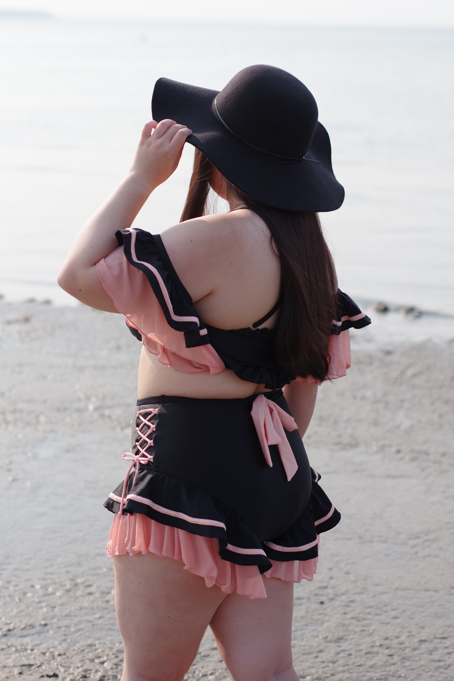 Back view of a woman wearing a whimsical black and pink ruffled swimsuit with lace-up details and a wide-brim hat at the beach.
