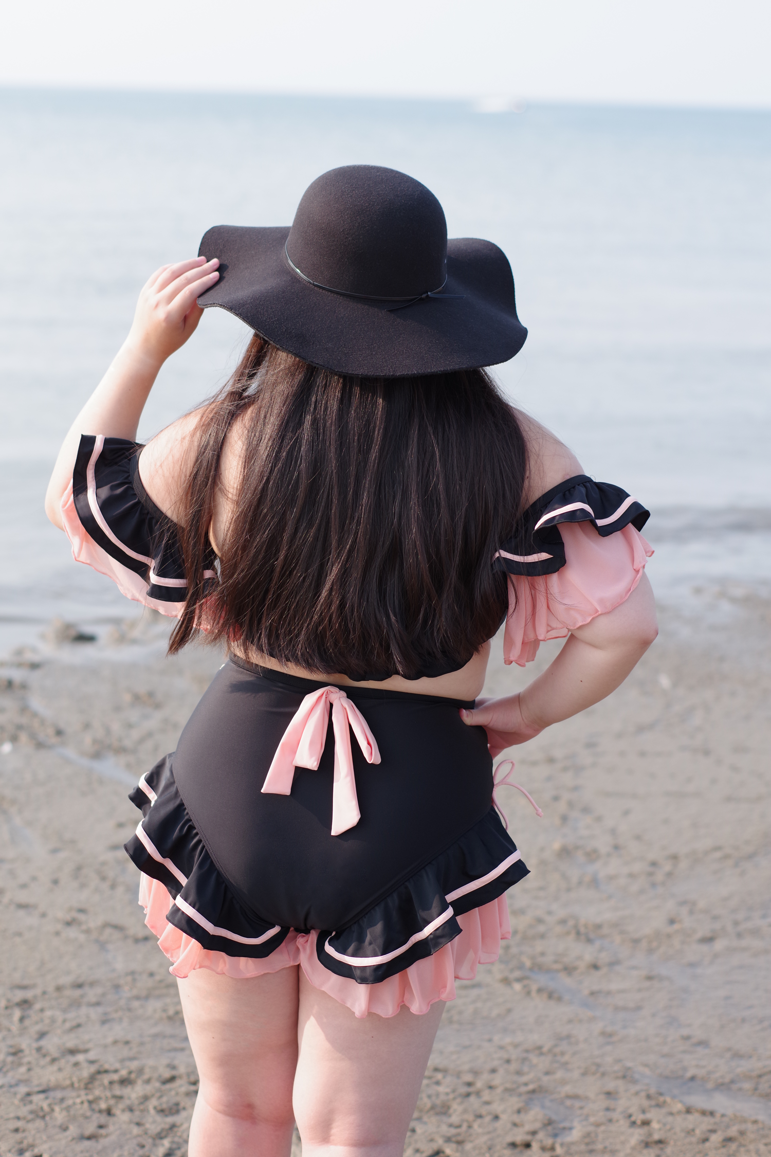 Back view of a woman wearing a whimsical black cat-inspired swimsuit with layered pink ruffles, bow tie details, and a wide-brim hat while standing on the beach.