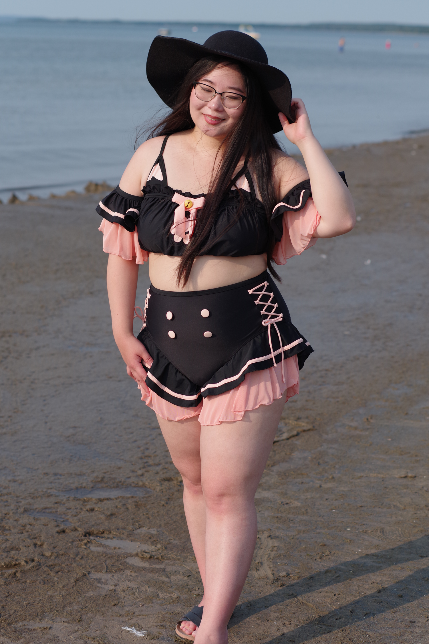 Woman wearing a whimsical black cat-inspired swimsuit with pink ruffles and lace-up details while walking along the beach in a wide-brim hat.