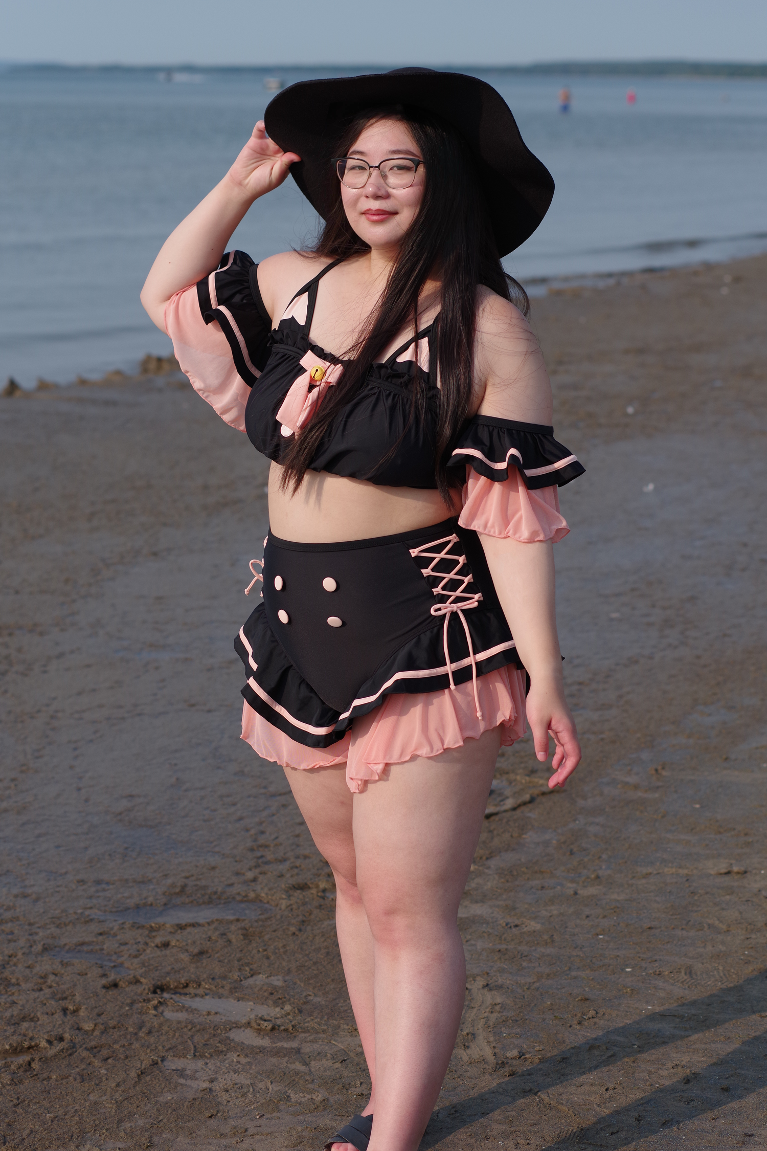Woman wearing a whimsical black cat-inspired swimsuit with pink ruffles, lace-up sides, and a wide-brim black sun hat while posing on the beach.