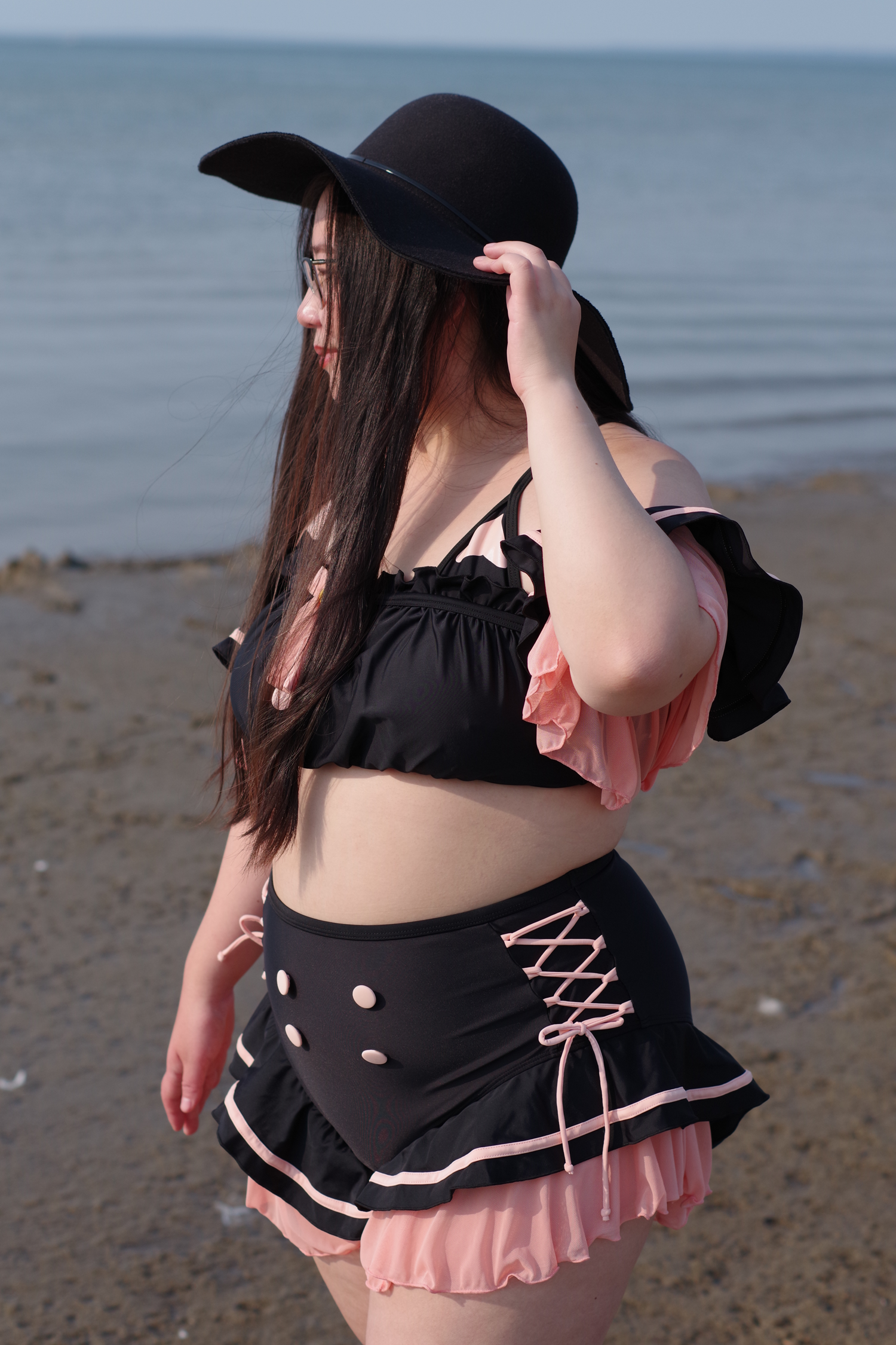 Woman wearing a whimsical black cat-inspired swimsuit with pink ruffle details, lace-up accents, and a wide-brim hat standing by the beach at sunset.