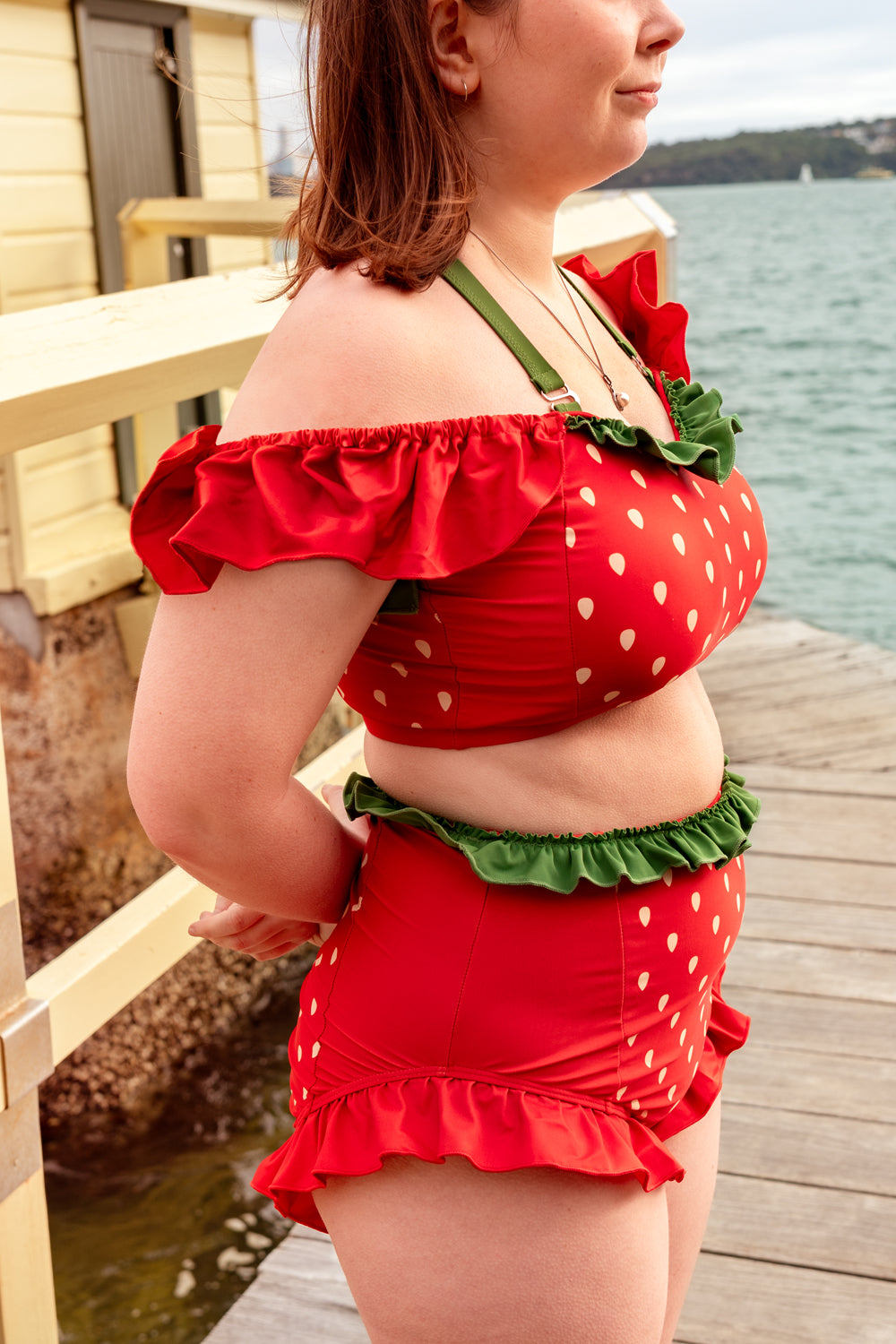 Side profile of a whimsical strawberry-inspired swimsuit with red ruffles, strawberry print, and green frill trim by the water.