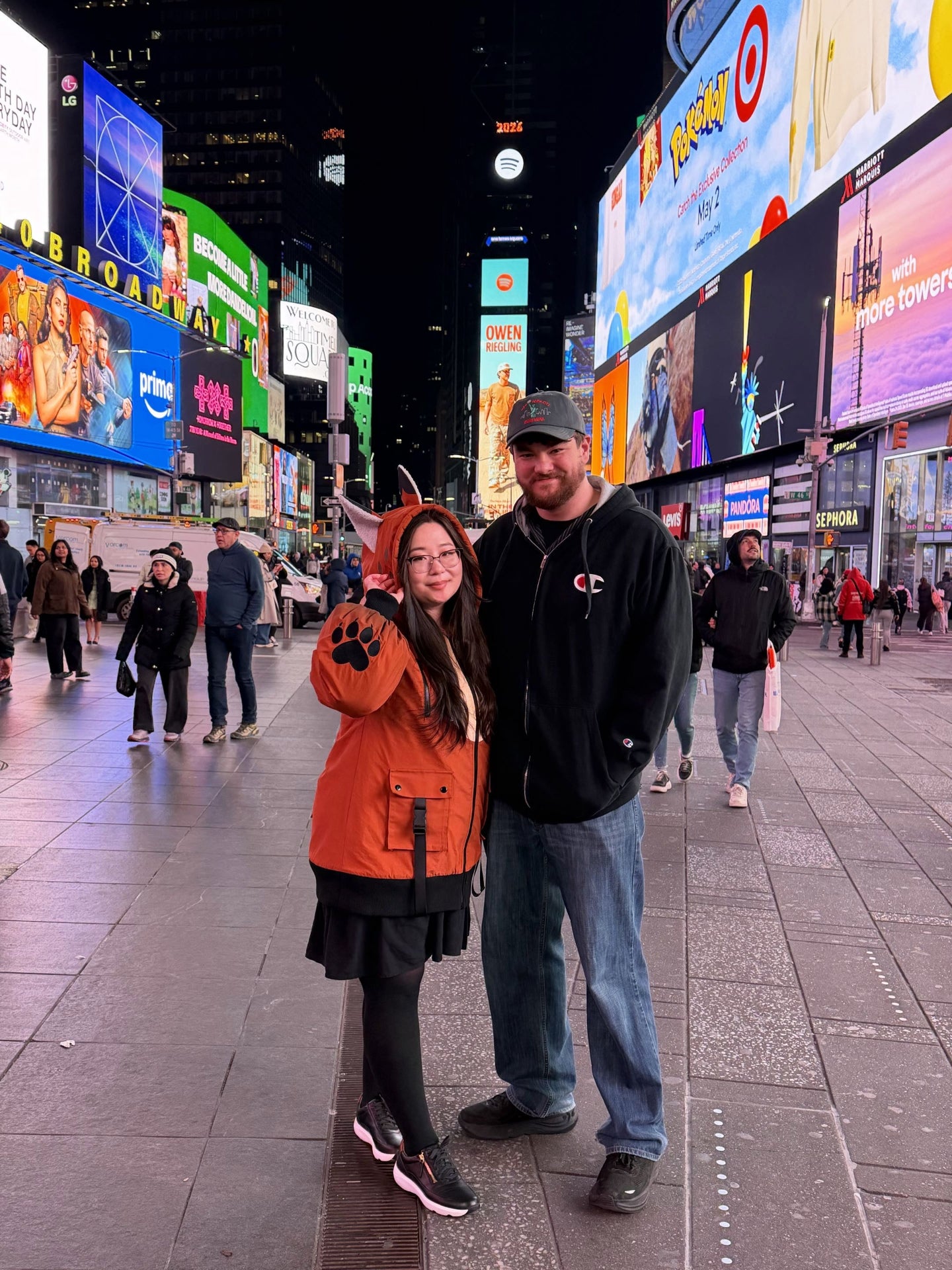Couple posing in Times Square at night with one person wearing a whimsical fox-inspired jacket with animal ears and paw print details.