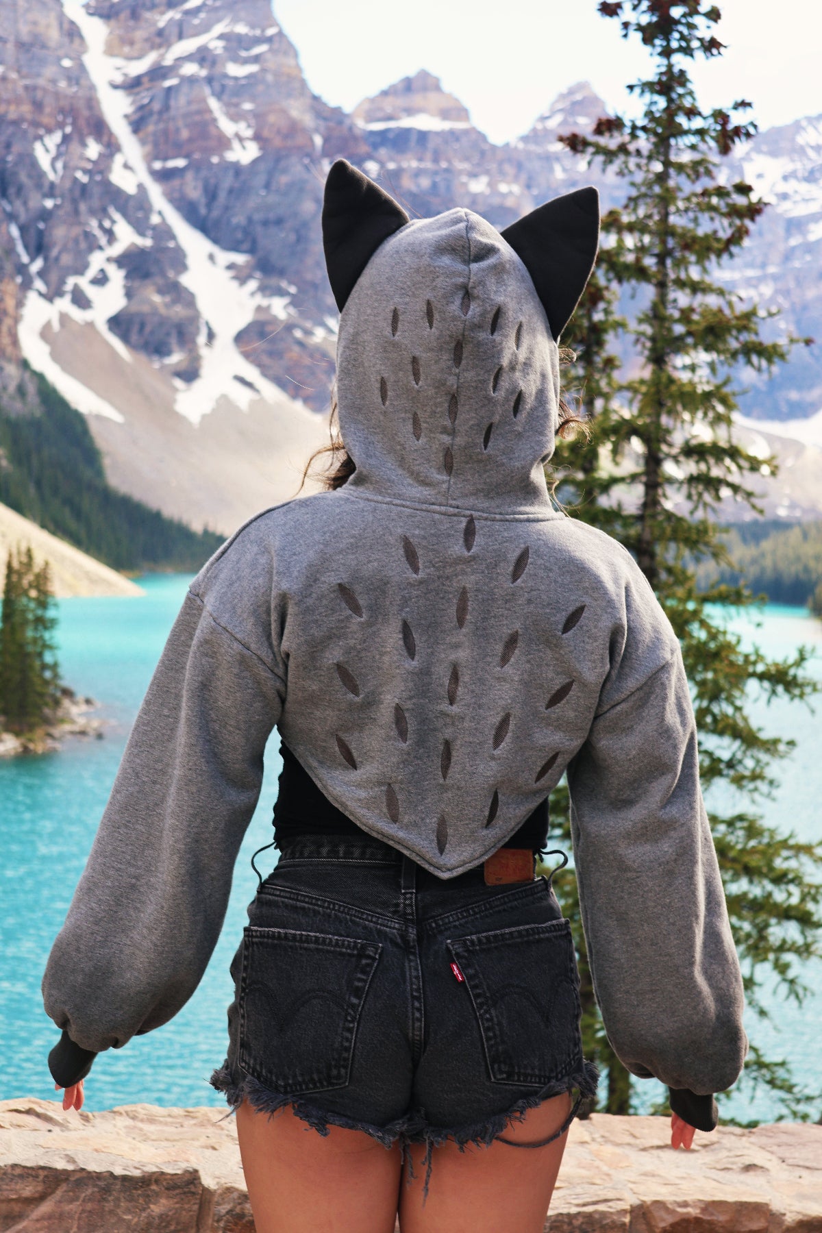 Back view of the Nyahallo Wolf Shrug featuring embroidered wolf fur detailing, wolf ears, and a cropped hoodie silhouette against a turquoise mountain lake.