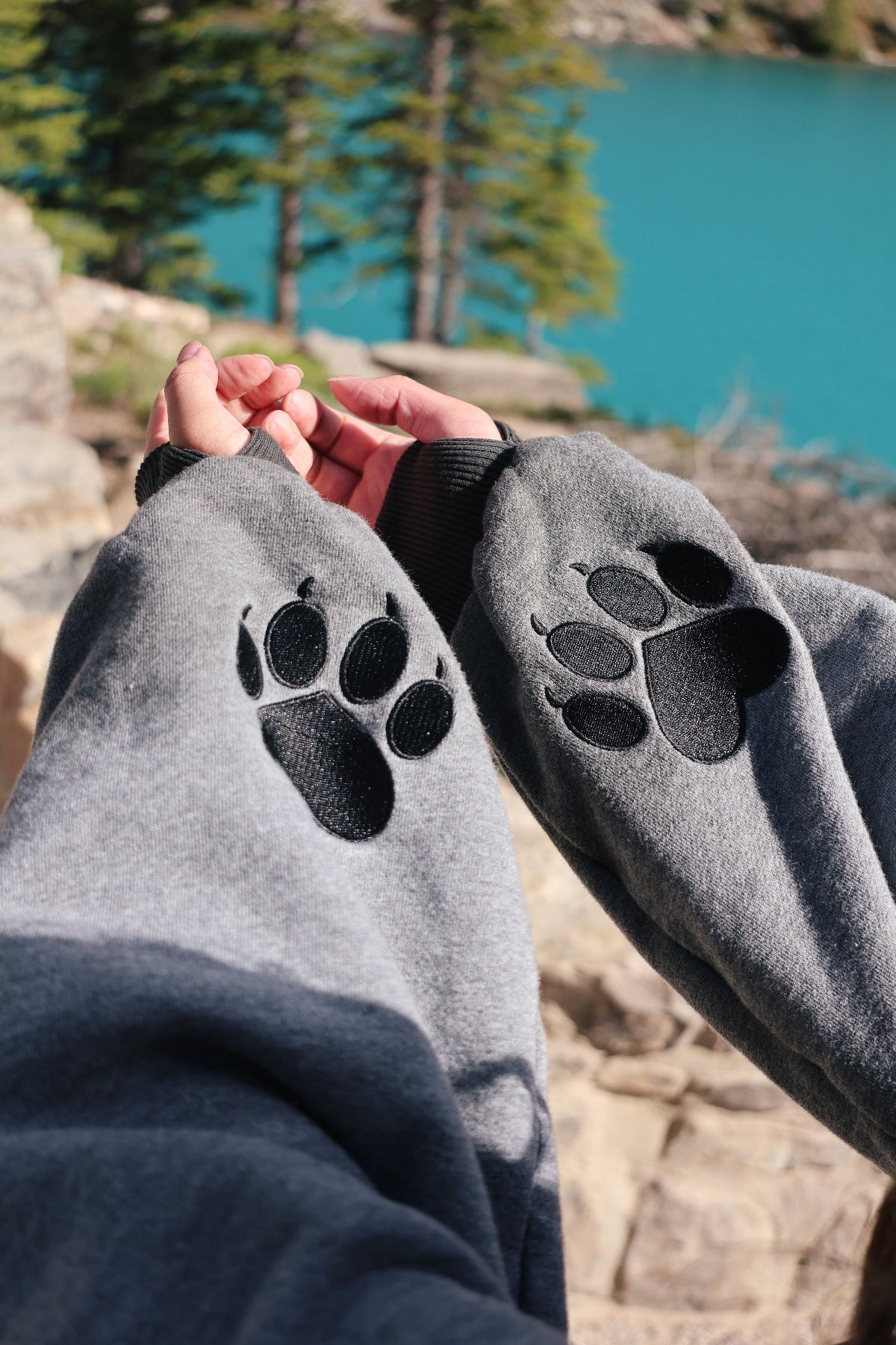 Close-up detail of the Nyahallo Wolf Shrug showcasing embroidered wolf paw prints on the sleeves with a turquoise mountain lake in the background.