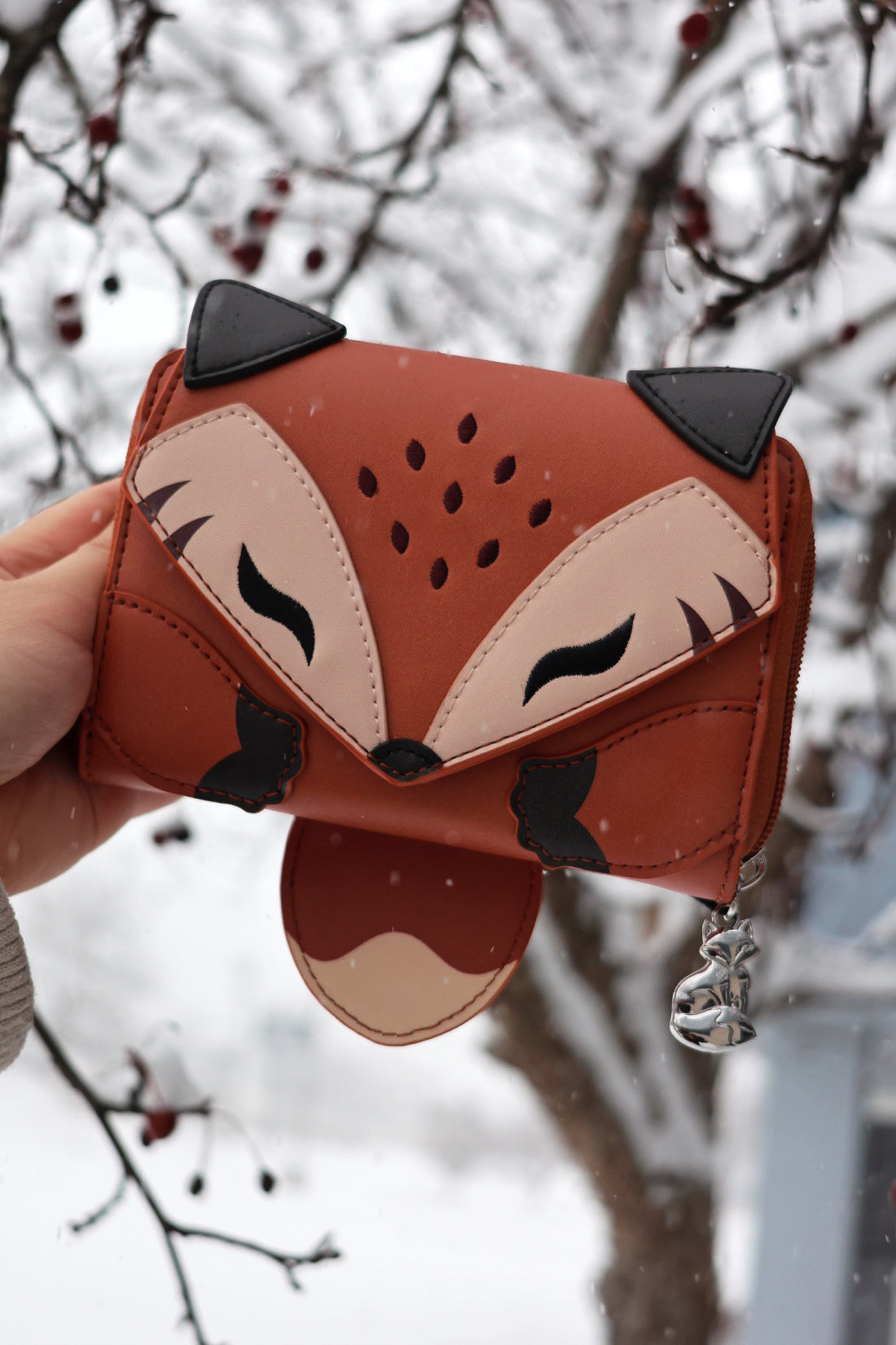 Woodland fox-shaped wallet with silver fox charm held outdoors in falling snow beside winter tree branches.