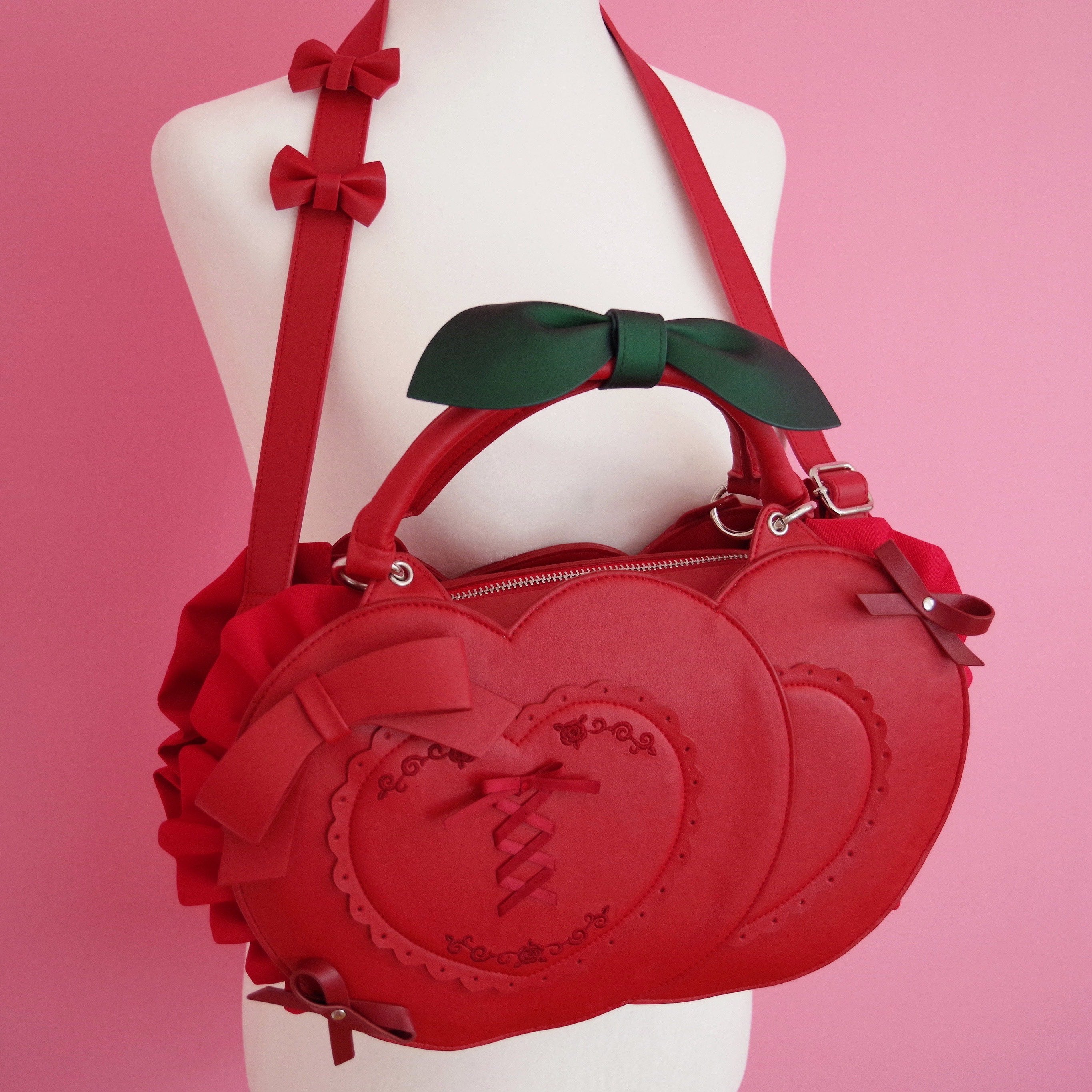 Cherry-inspired heart-shaped handbag featuring layered heart panels, decorative bows, leaf handle accents, and adjustable straps displayed on a mannequin against a pink background.