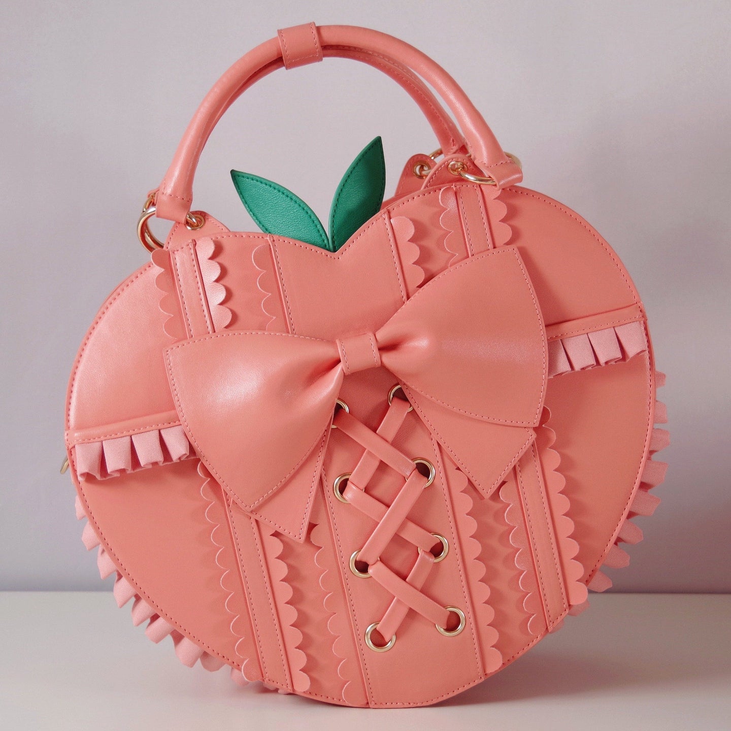 Peach-shaped handbag with oversized bow, lace-up corset details, ruffled trim, green leaf accents, and top handles from the Frilly Fruits collection.