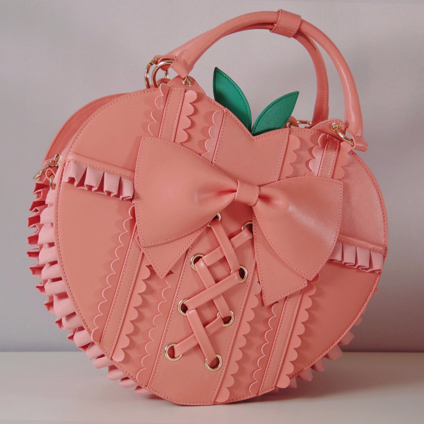 Peach-shaped handbag with oversized bow, corset lacing, scalloped trim, ruffled sides, and green leaf accents viewed from an angle.