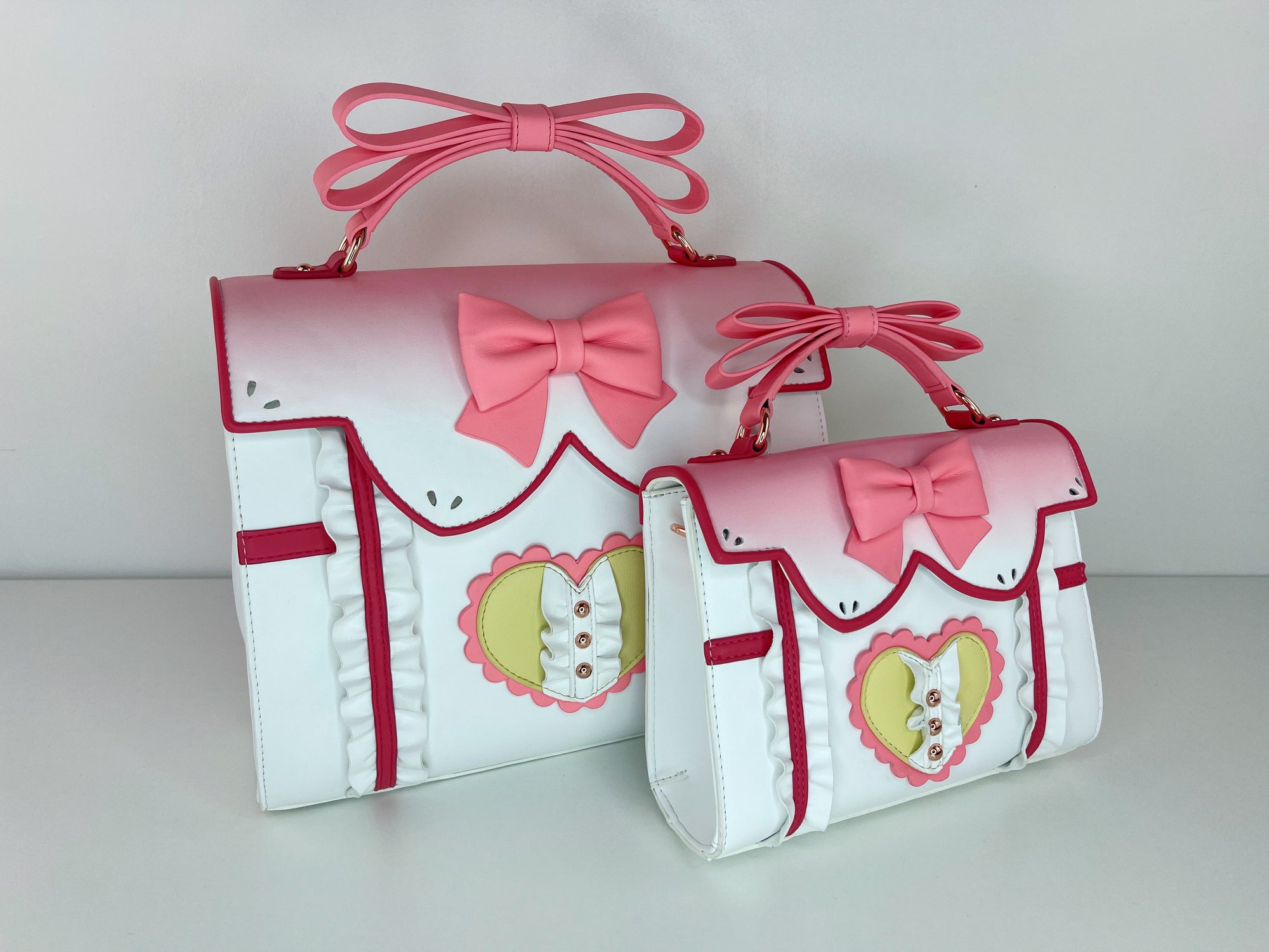 Two Madoka-inspired satchel bags in large and small sizes, featuring pink bows, scalloped collars, heart embellishments, and structured top handles. Displayed against a plain white background.