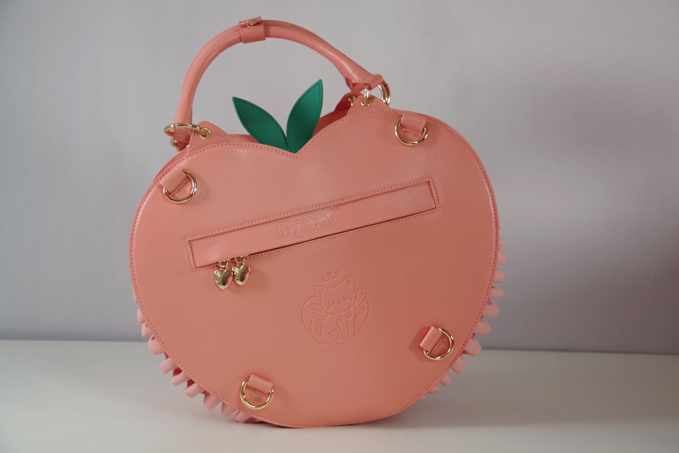 Back view of peach-shaped handbag featuring rear zipper pocket, detachable backpack straps, gold hardware, and leaf accents.